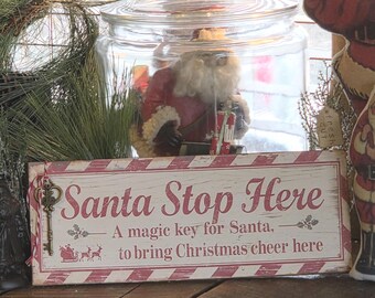 May include: A rectangular wooden sign with a white wood grain finish and red striped borders. The text reads Santa Stop Here, A magic key for Santa, to bring Christmas cheer here. A metal skeleton key is attached to the left side with twine.