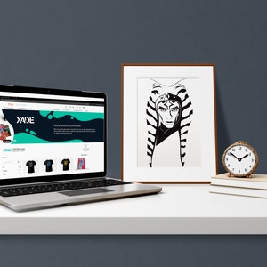 May include: A laptop computer displaying a website with the text "XADE" and a black and green graphic design. The website features a selection of t-shirts with different designs. A framed black and white illustration of a person with a headdress is on the right side of the laptop. A small gold clock is on a stack of books.