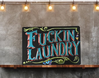 May include: A black sign with a white border and gold accents. The sign says "Fuckin Laundry" in a stylized font with blue and red lettering.