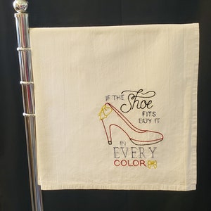 May include: White kitchen towel with red embroidered high heel shoe and the text "If the shoe fits buy it in every color" with yellow embroidered bows.