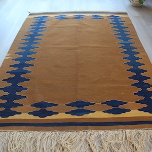 May include: A brown rug with a blue and yellow geometric pattern. The rug has a fringe border.