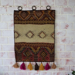 Puede incluir: Un tapiz tejido de pared marrón y beige con patrones geométricos y borlas. El tapiz está colgado en una pared de ladrillo blanco.
