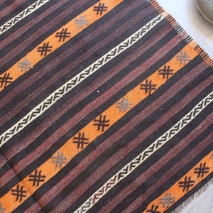 May include: A brown and orange striped rug with a geometric pattern. The rug has a fringe border.