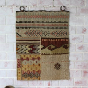Puede incluir: Un tapiz de pared rectangular de patchwork con varios patrones geométricos en tonos beige, marrón, naranja y rojo. Tiene dos anillos metálicos en la parte superior para colgarlo. El fondo es una pared de ladrillos blancos.