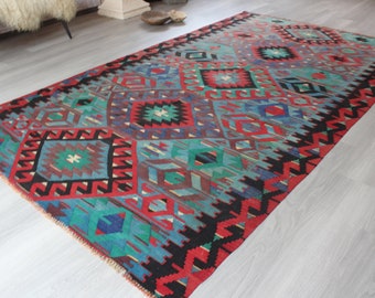 May include: A traditional Turkish rug with a geometric pattern in shades of red, blue, green, and black. The rug has a fringed edge and is woven with intricate details.