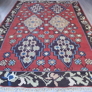 May include: A traditional red, blue, and beige patterned rug with a geometric design. The rug has a dark brown border with floral motifs.