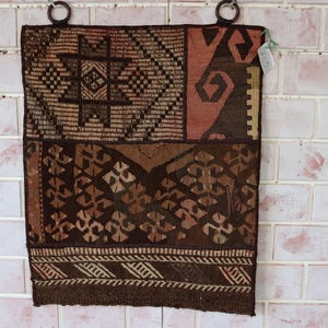 May include: A brown and beige woven tapestry with geometric and floral patterns. The tapestry is hanging on a white tiled wall.