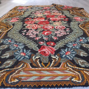May include: A vintage floral rug with a dark background and a central design of red, pink, and white flowers. The rug has a brown, gold, and blue border with a geometric pattern.