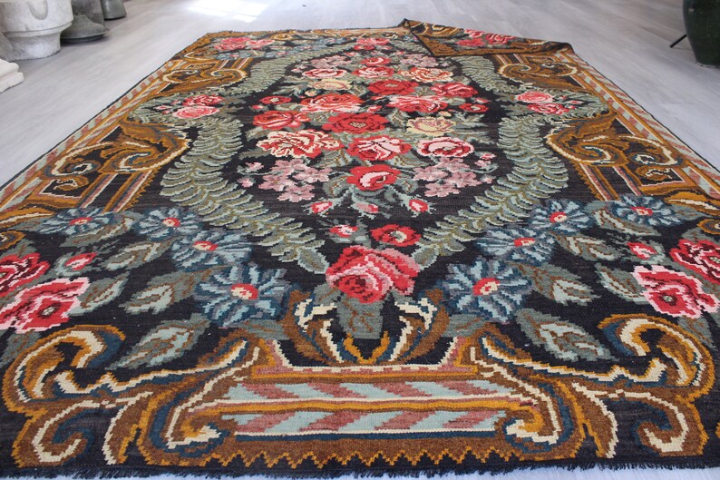 May include: A vintage floral rug with a dark background and a central design of red, pink, and white flowers. The rug has a brown, gold, and blue border with a geometric pattern.