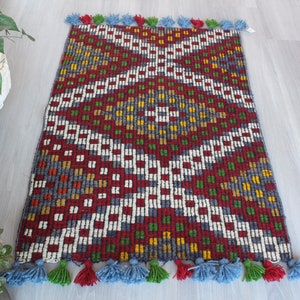 May include: A colorful geometric patterned rug with a red, white, green, blue, and yellow color scheme. The rug has a fringe border with tassels in red, blue, and green.