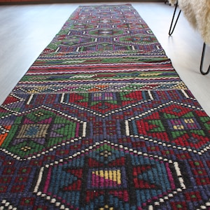May include: A long, narrow rug with a colorful geometric pattern. The rug is woven with a variety of colors, including red, green, blue, and yellow. The pattern is intricate and detailed, and the rug is made of a soft, durable material.