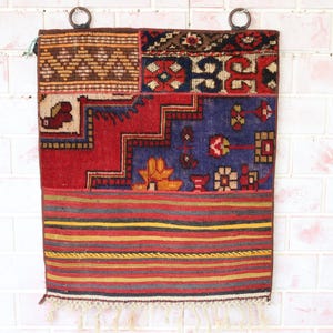 Op de afbeelding: Een kleurrijk patchwork tapijt met een rood, blauw en geel patroon. Het tapijt hangt aan een witte bakstenen muur en heeft franjes aan de onderkant.