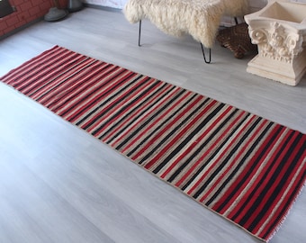 Può includere: Un tappeto lungo e stretto a strisce rosse, nere e crema. Il tappeto è realizzato in materiale tessuto e ha un bordo con frange.