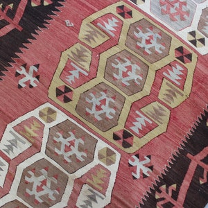 Może przedstawiać: Antyczny turecki dywan kilim z geometrycznym wzorem w odcieniach czerwieni, brązu, beżu i bieli. Dywan charakteryzuje się powtarzającym się wzorem sześciokąt&oacute;w i tr&oacute;jkąt&oacute;w.