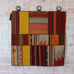 Puede incluir: Una alfombra de patchwork colorida con patrones geométricos en tonos rojos, naranjas, amarillos, marrones y blancos. La alfombra está colgada en una pared de ladrillo blanco con tres ganchos metálicos.