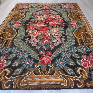May include: A vintage floral rug with a dark background and intricate patterns. The rug features a central design of red, pink, and white flowers surrounded by green leaves and brown vines. The border of the rug is a rich brown with gold accents.