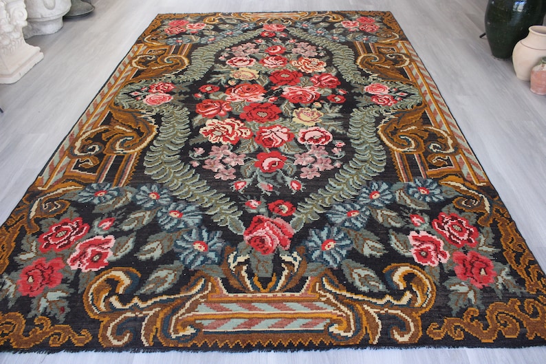 May include: A vintage floral rug with a dark background and intricate patterns. The rug features a central design of red, pink, and white flowers surrounded by green leaves and brown vines. The border of the rug is a rich brown with gold accents.