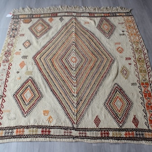 May include: A white woven rug with a diamond pattern in shades of red, orange, brown, and black. The rug has a fringe border.