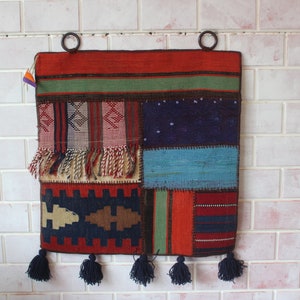 以下が含まれることがあります： 赤、青、緑、茶色など、さまざまな幾何学模様と色が特徴の、パッチワークデザインのカラフルなタペストリー壁掛け。タペストリーは下部にフリンジがあり、2つの金属製のリングで吊り下げられています。