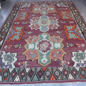 May include: A large, rectangular rug with a deep burgundy background and intricate geometric patterns in shades of orange, teal, and cream. The rug features a detailed border and a central medallion design, showcasing a traditional style.