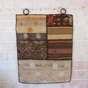 以下が含まれることがあります： 茶色、オレンジ、ベージュの色合いの幾何学模様が様々なパッチワークの敷物。敷物は白いレンガの壁に、上部に金属製のリングが2つ付いて吊り下げられている。