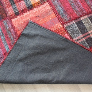 K&ouml;nnte beinhalten: Ein roter, blauer und grauer Patchwork-Teppich mit einer dunkelgrauen R&uuml;ckseite. Der Teppich besteht aus verschiedenen Stoffen und hat einen Fransenrand.