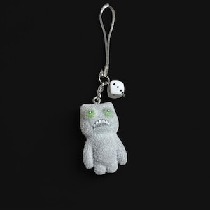 Peut inclure: Un porte-clés argenté avec une petite figurine grise de style cartoon, aux yeux verts et à la bouche renfrognée. Un dé blanc à points noirs est suspendu à côté. Le fond est noir uni.