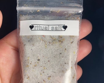 May include: A small clear plastic bag filled with white salt and small brown and green herbs. The bag has a white label with black text that reads "ritual salt" and two black hearts.
