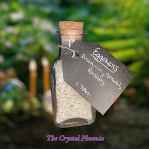 May include: A small glass jar filled with crushed eggshells. A black tag is attached to the jar with the text "Eggshells Protection, renewal, fertility 1.7 oz."