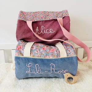 Könnte beinhalten: Zwei Cordtaschen, eine magenta und eine blaue, mit floral gemusterten Oberteilen. Auf der magentafarbenen Tasche ist "Alice" und auf der blauen Tasche "Lili-Rose" gestickt. Ein rosa Gurt und ein Holzspielzeug in Vogelform sind ebenfalls zu sehen.