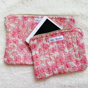 Peut inclure: Deux étuis pour tablette en tissu matelassé avec un motif floral rose et beige. Un étui contient une tablette. Les étuis ont une fermeture éclair et une étiquette "Lili Broderie".