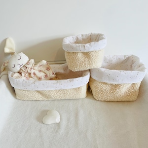 Peut inclure: Trois paniers de rangement en tissu sherpa couleur crème avec des doublures en mousseline blanche à motif d'étoiles dorées. Un panier rectangulaire contient un lapin en peluche à motif floral, et un petit objet blanc en forme de cœur est posé devant.