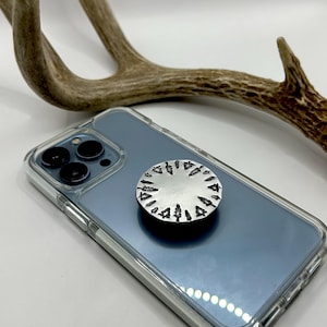 Op de afbeelding: Een zilveren telefoon pop socket met een ontwerp van zwarte bomen rond de rand.