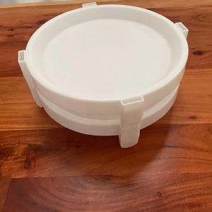 Peut inclure: Un plateau circulaire en plastique blanc doté d'un rebord surélevé et de quatre supports verticaux régulièrement espacés s'étendant de la base au bord supérieur, créant une structure empilable avec une surface intérieure lisse et plate.