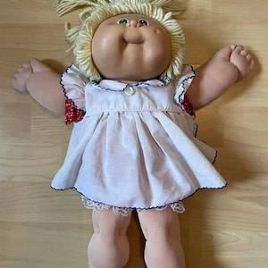 Puede incluir: Una muñeca de piel clara, ojos verdes y cabello de lana rubia recogido en una coleta. La muñeca viste un vestido blanco con cuello de volantes, un botón y mangas con estampado floral rojo, junto con unos bombachos blancos con encaje.
