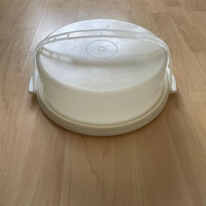 Może przedstawiać: Biały plastikowy pojemnik na ciasto z przezroczystym uchwytem i pokrywką. Pojemnik jest okrągły z podstawą i kopulastym wierzchem, przeznaczony do ochrony i transportu wypieków. Uchwyt jest wygięty dla łatwego przenoszenia.