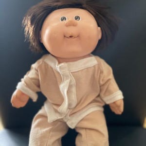 Könnte beinhalten: Eine Cabbage Patch Kid Puppe mit braunen Haaren, die einen hellbraunen Schlafanzug mit weißen Verzierungen trägt.