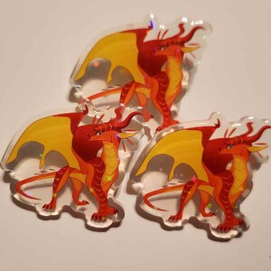 Puede incluir: Tres pines de drag&oacute;n de acr&iacute;lico transparente. Cada pin presenta un drag&oacute;n rojo y naranja con alas amarillas. Los dragones est&aacute;n de pie con las alas extendidas. Los pines tienen un efecto de brillo hologr&aacute;fico.