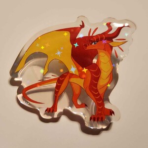 Puede incluir: Una pegatina hologr&aacute;fica transparente con un drag&oacute;n de dibujos animados en tonos rojos, naranjas y amarillos. El drag&oacute;n tiene grandes alas y brillos en forma de estrella. La pegatina est&aacute; cortada en forma de nube.