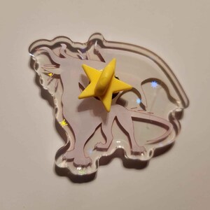 Puede incluir: Un pin de acr&iacute;lico transparente con un dise&ntilde;o de drag&oacute;n morado claro y un pin de estrella amarilla. El drag&oacute;n tiene alas y detalles de estrellas. El pin es un accesorio &uacute;nico.