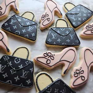 Könnte beinhalten: Eine Sammlung von neun Zuckerkeksen, die mit schwarzen und weißen Louis-Vuitton-inspirierten Mustern und rosa High Heels verziert sind. Die Kekse befinden sich auf einer weißen Marmorfläche.