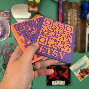 May include: Two orange and purple 3D printed coasters with a QR code and the word "ETSY" on them.