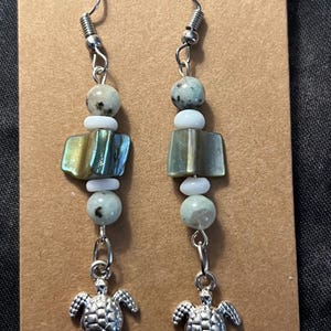 May include: A pair of dangle earrings with silver-tone hooks. Each earring features a combination of beads in shades of green, white, and speckled gray, along with a rectangular iridescent bead. The earrings are finished with a small, detailed silver turtle charm.