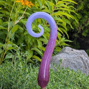 May include: A decorative garden stake with a purple and lavender swirl design. The stake is made of glass and has a bronze-colored metal base. The stake is placed in a garden with green foliage and yellow flowers.