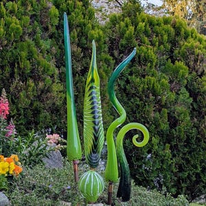 May include: A collection of vibrant green glass garden art sculptures. The pieces include tall, slender spires, a striped column, and a curled design, all set against a backdrop of lush green foliage and colorful flowers. The sculptures are mounted on metal rods.