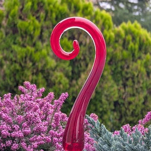 May include: A vibrant red glass garden stake with a curled top. The stake is set against a backdrop of green and purple foliage, creating a striking contrast. The stake is mounted on a copper-colored base.