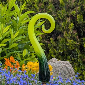 May include: A decorative garden stake with a swirling, green glass top that fades to dark green at the base. The stake is set in a bed of blue flowers, with orange and yellow flowers in the background.