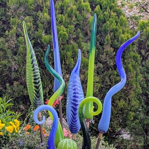 May include: A collection of colorful glass garden art sculptures. The pieces feature various shapes, including tall, slender forms and curved, swirling designs. The color palette includes shades of blue, green, and a touch of yellow, creating a vibrant display in a garden setting.