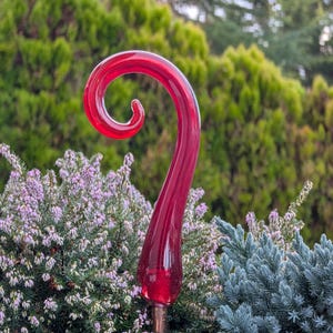 May include: A vibrant red glass garden stake with a curled top. The stake is set against a backdrop of green and purple foliage, adding a pop of color to any outdoor space. The glass has a glossy finish.