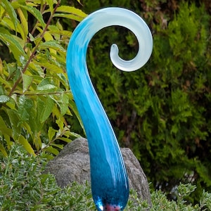 May include: A blue and white glass garden stake with a curved, spiral design. The stake transitions from a deep blue to a light blue and white swirl. The stake is mounted on a copper-colored metal rod, and is surrounded by green foliage.